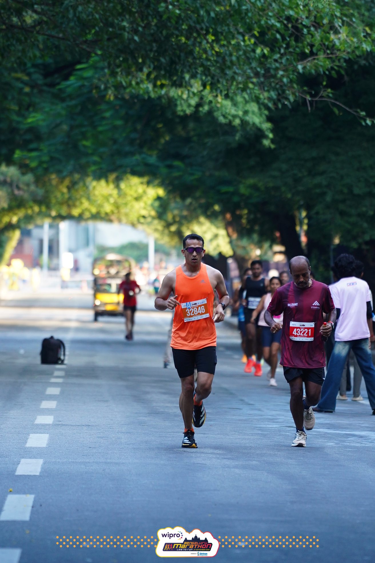 Anand runs down the middle of a road under a thick green tree canopy, other runners behind him on the right.