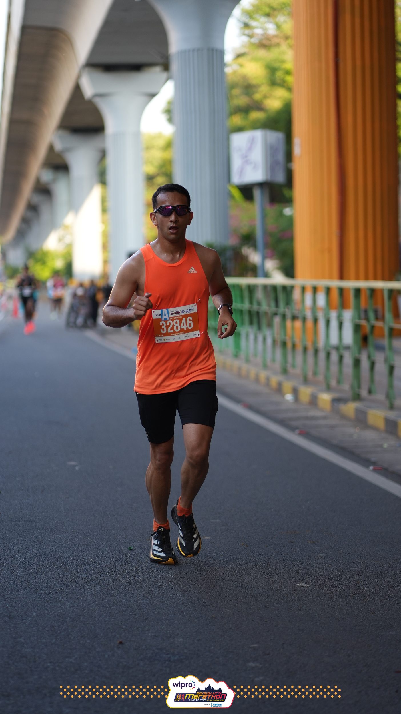 Anand, bib 32846, in an orange singlet and mirrored sunglasses, running alone under the elevated metro line, orange and grey pillars behind him.