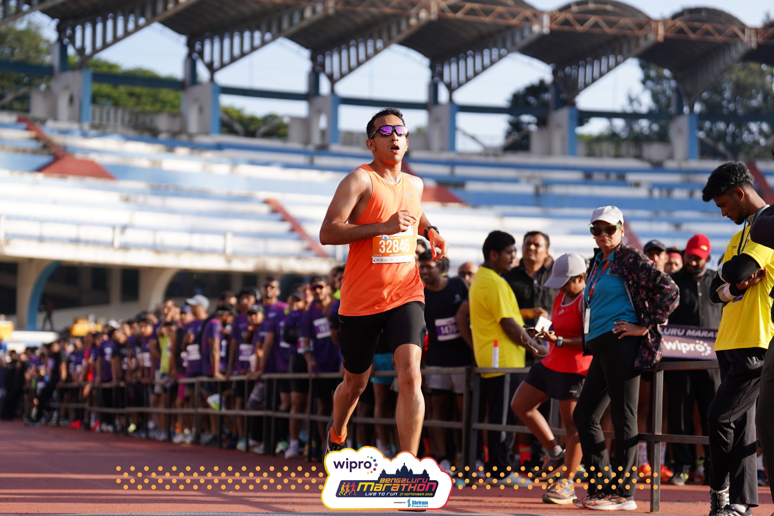 Anand runs along the red track inside Sree Kanteerava Stadium, spectators and runners lined up along the barriers beside him and the stands behind.