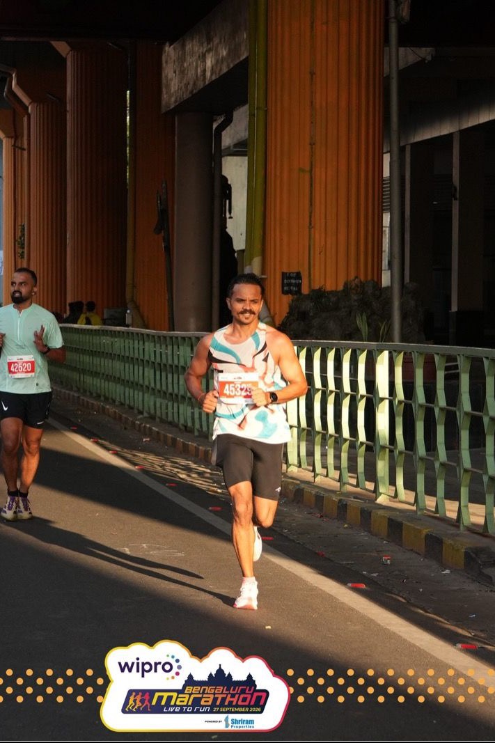 Uttam runs in morning sunlight under the elevated metro line, orange pillars behind him and another runner to his right.