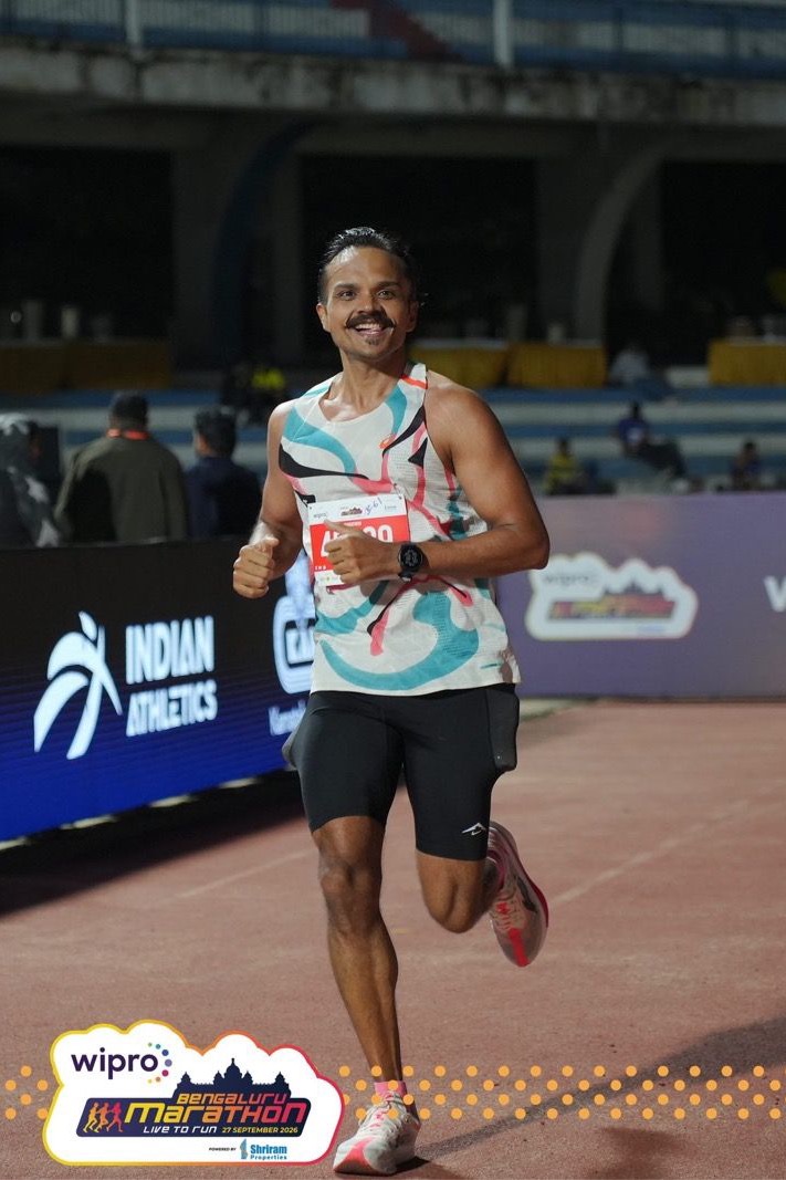 Uttam smiles and gives a thumbs-up as he runs along the track past an Indian Athletics board.