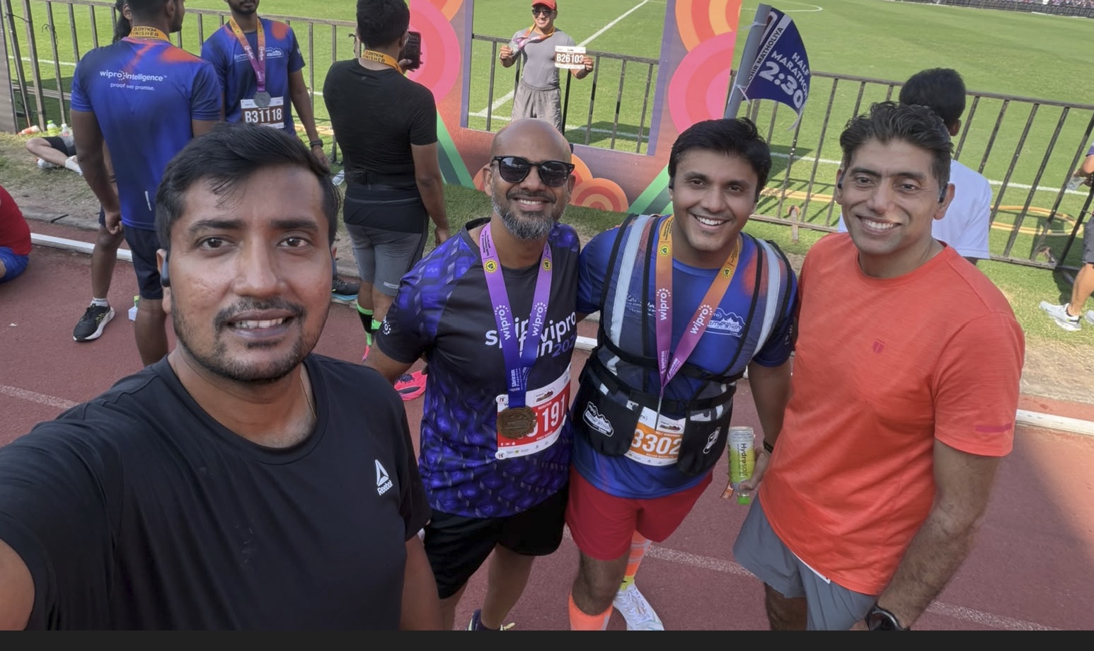 A selfie of four runners on the stadium track after the race, two wearing finisher medals. Vineet is on the right in an orange T-shirt, with the football pitch and a half marathon 2:30 pacer flag behind them.