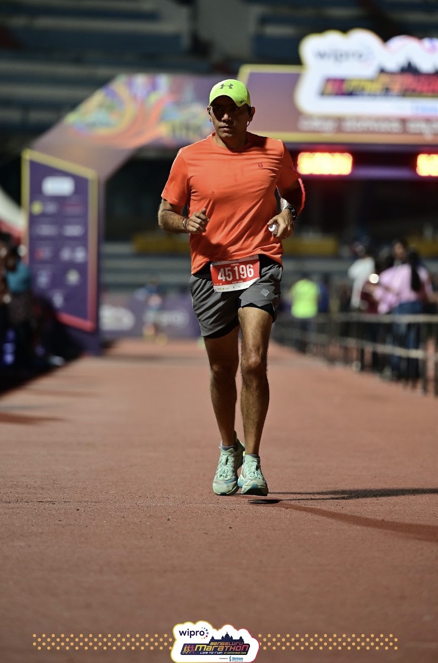 Vineet, bib 45196, in an orange T-shirt, grey shorts and a yellow cap, runs along the red track inside Sree Kanteerava Stadium with the finish arch behind him.