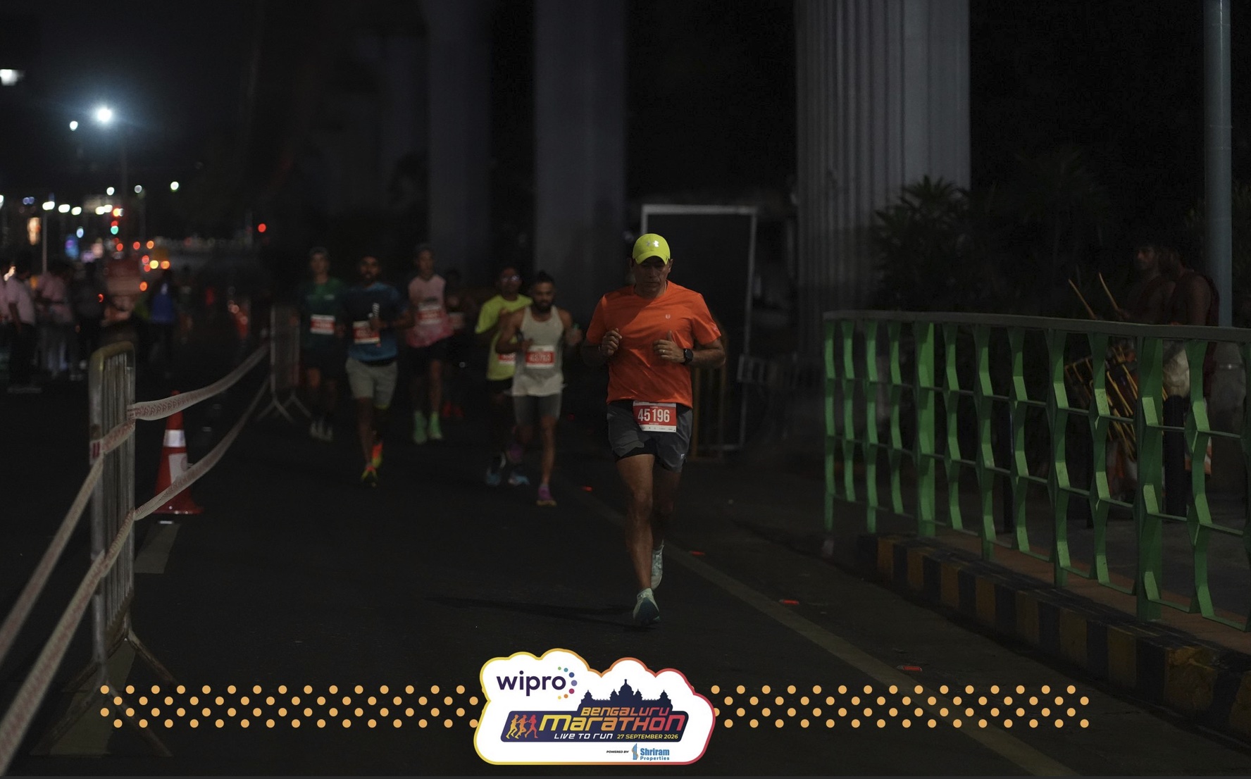 Vineet, bib 45196, in an orange T-shirt and yellow cap, runs in the dark under the metro line ahead of a group of runners.