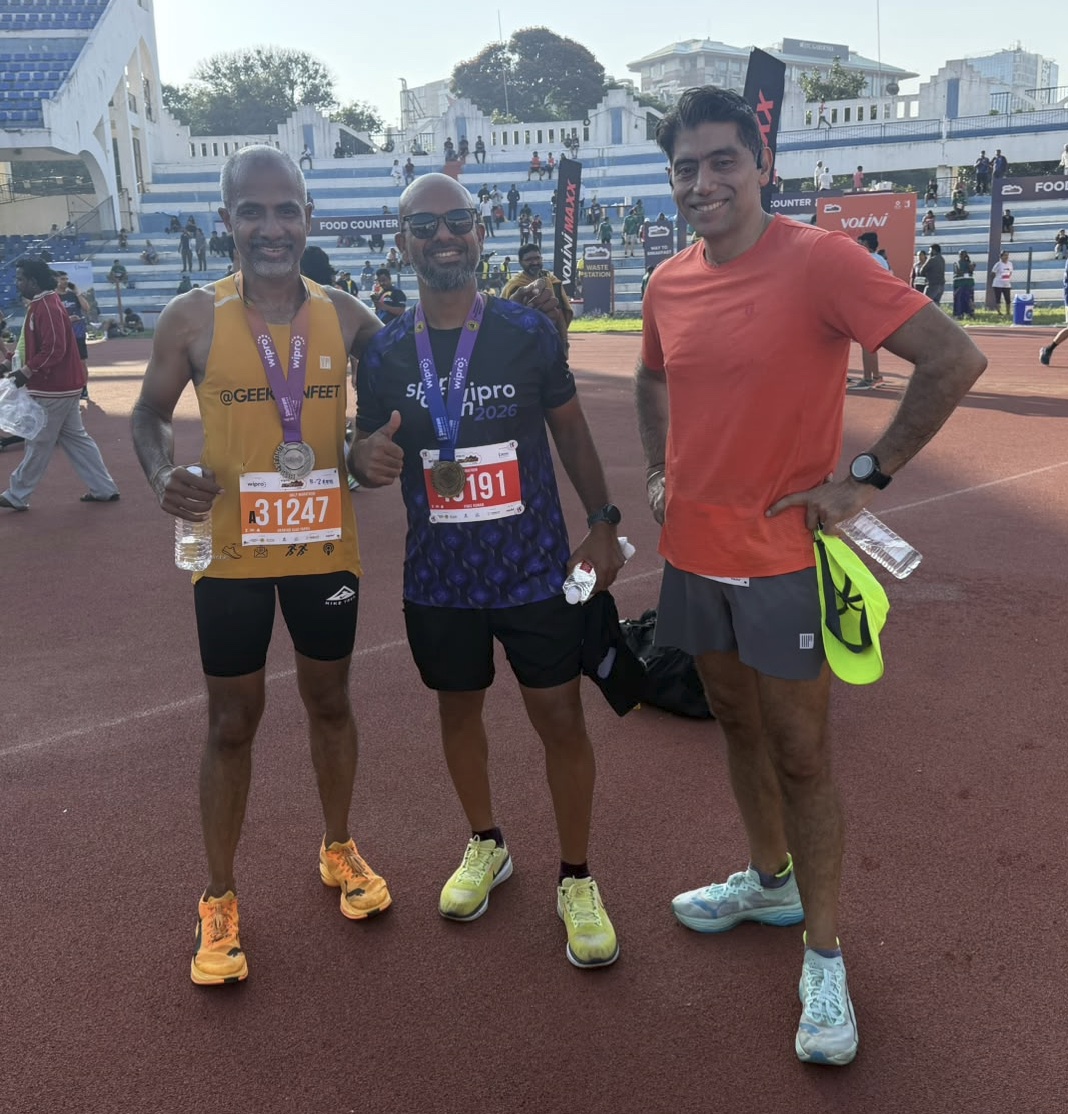 Three runners stand on the stadium track after the race, two wearing finisher medals. Vineet, on the right in an orange T-shirt, holds his yellow cap and a water bottle.