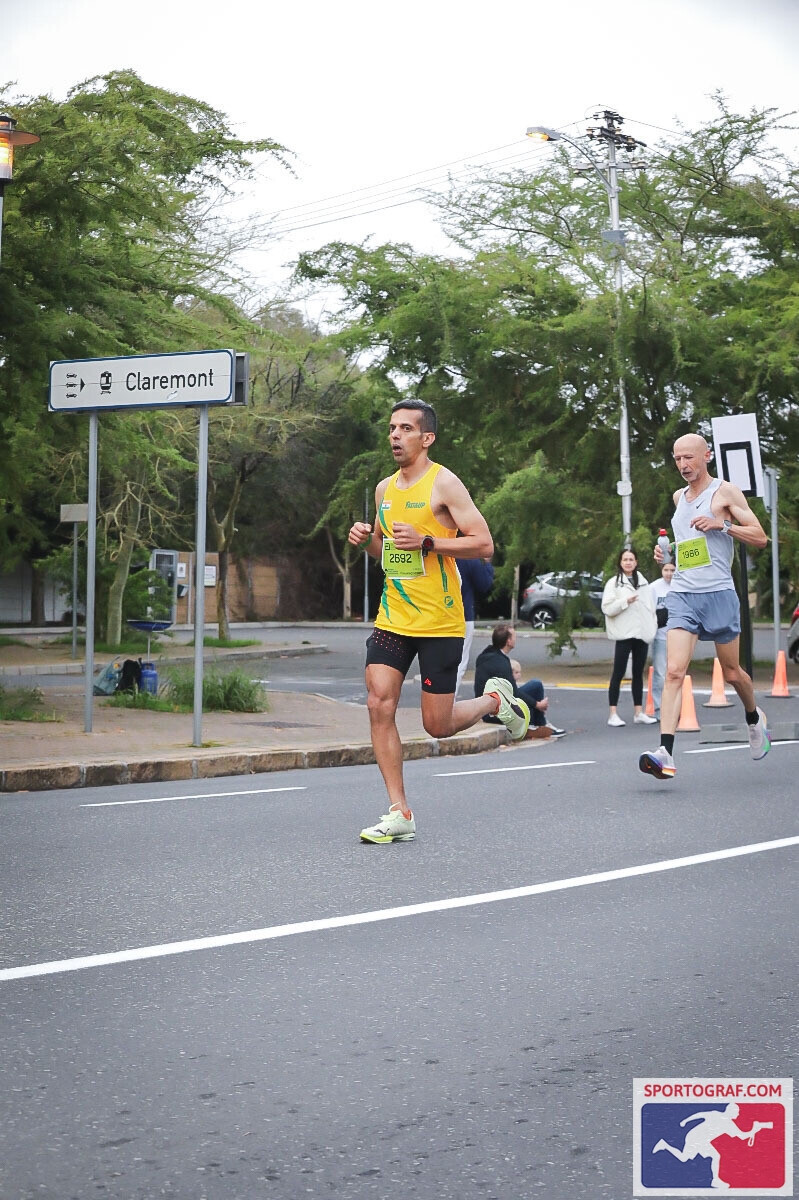 A road sign reads 'Claremont' as Vijay strides past a fellow runner on a tree-lined Cape Town street.