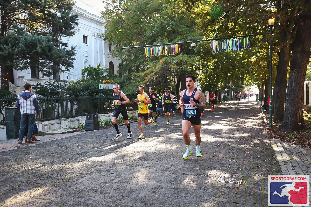 Three runners stride down a long cobblestoned street past a white church facade, festive bunting strung overhead, spectators lining the pavement.