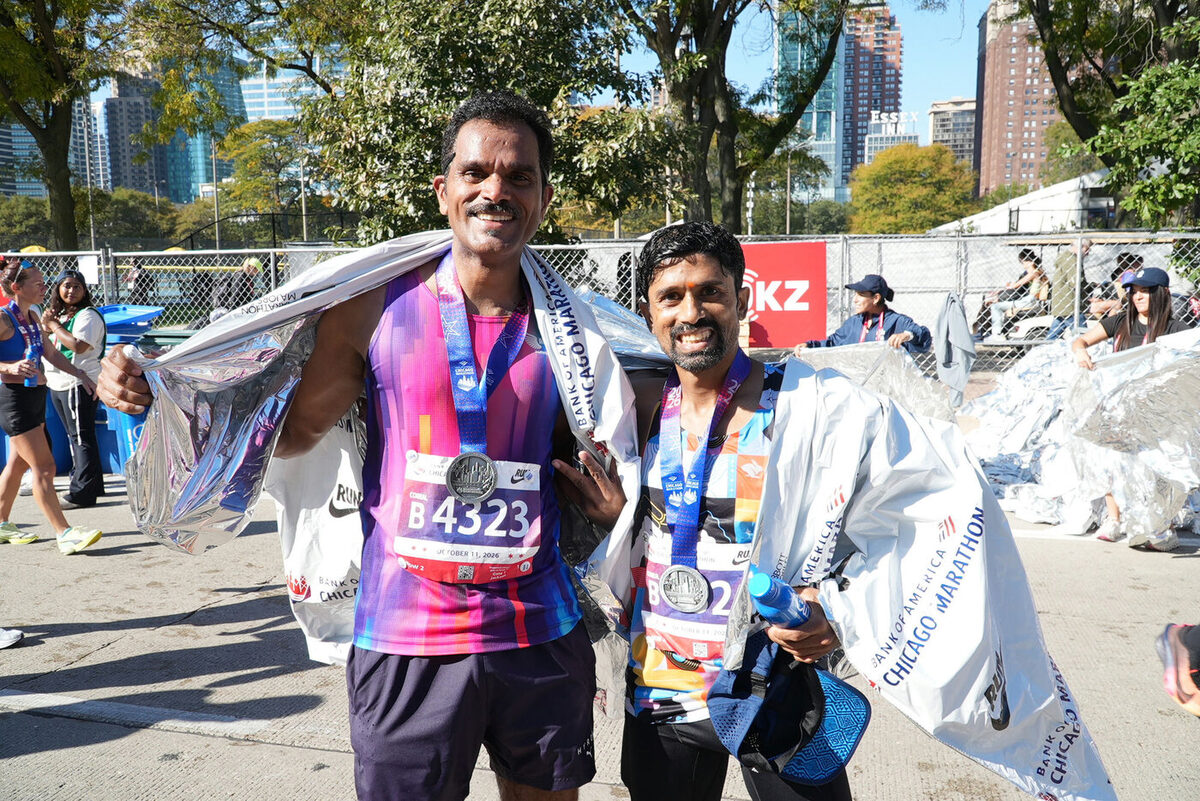 Murali Gaddam and Vikram stand wrapped in silver foil blankets after the Chicago Marathon, medals around their necks, the city skyline behind them.