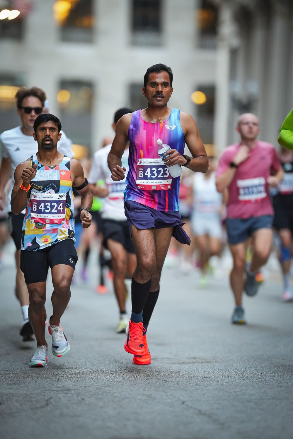 Murali Gaddam, bib B4323, and Vikram, bib B4324, run shoulder to shoulder through a Chicago street, Murali carrying a water bottle.