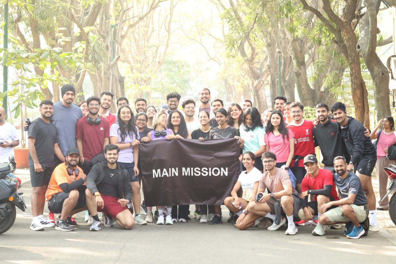 A large group of young runners holds a black banner reading 'MAIN MISSION' at the end of a Sunday morning run in Bengaluru.