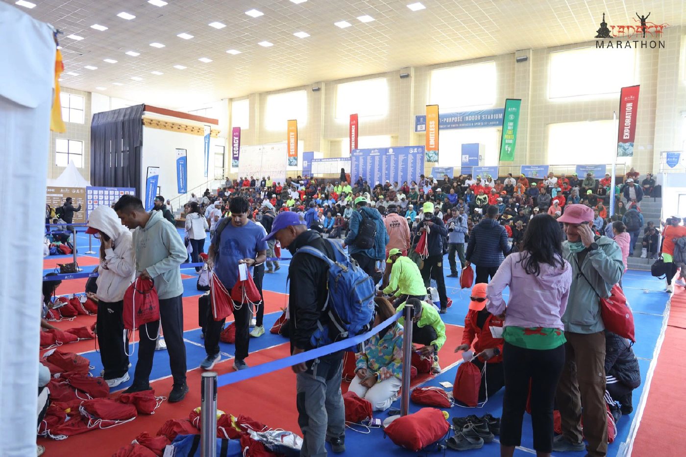 A large indoor hall packed with runners collecting kit and red drop bags before the Ladakh Marathon, with race banners along the walls.