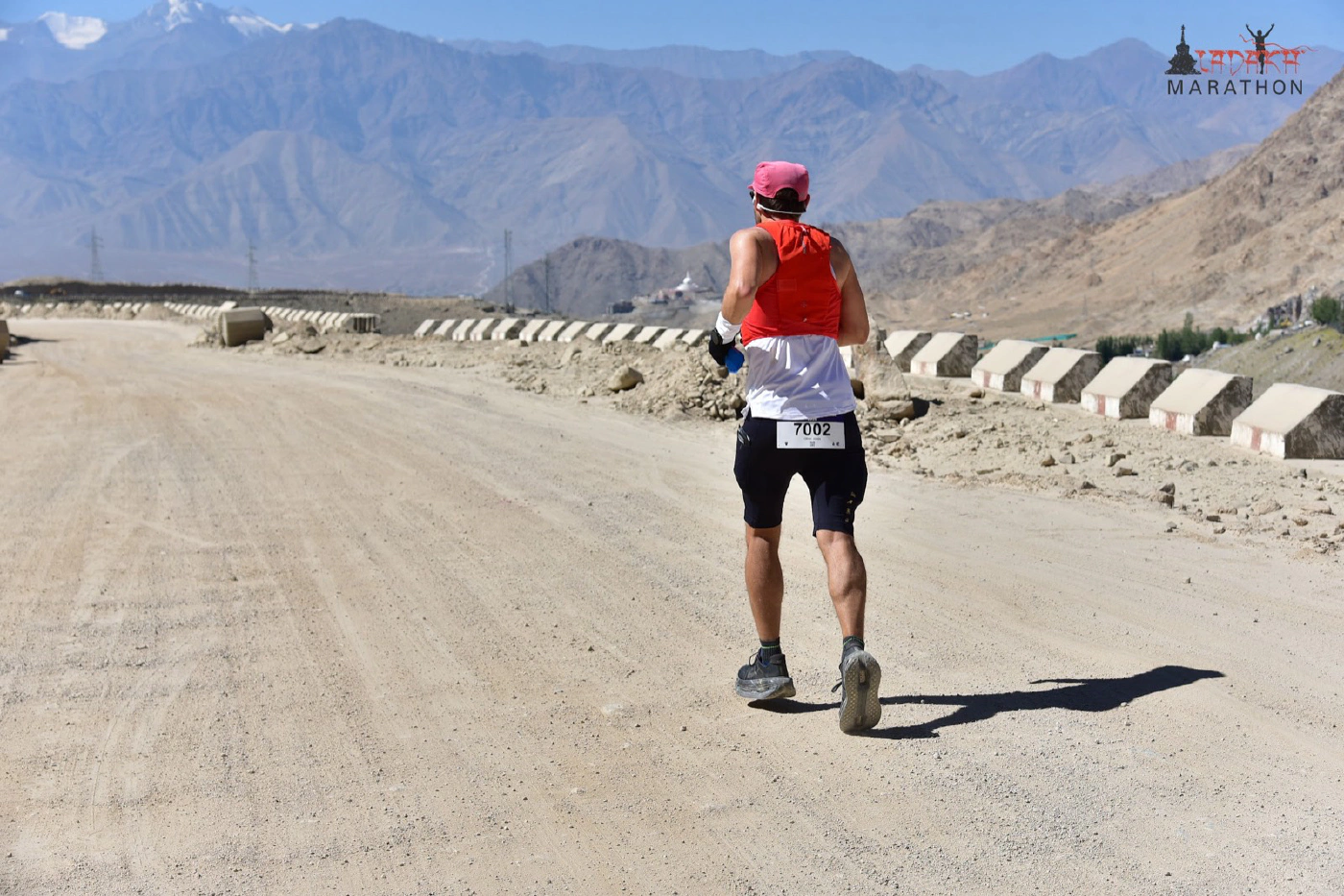 Oliver running on a high gravel road with the town of Leh visible far below in the valley, mountains stretching beyond.