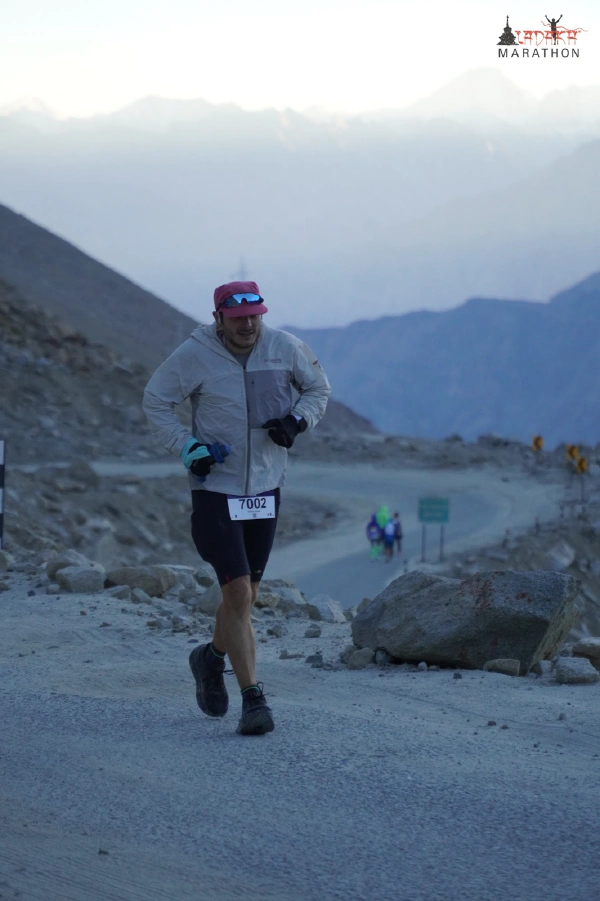 Oliver running on a gravel mountain road in the early morning half-light, wearing a windcheater, cap and gloves, with other runners behind him.
