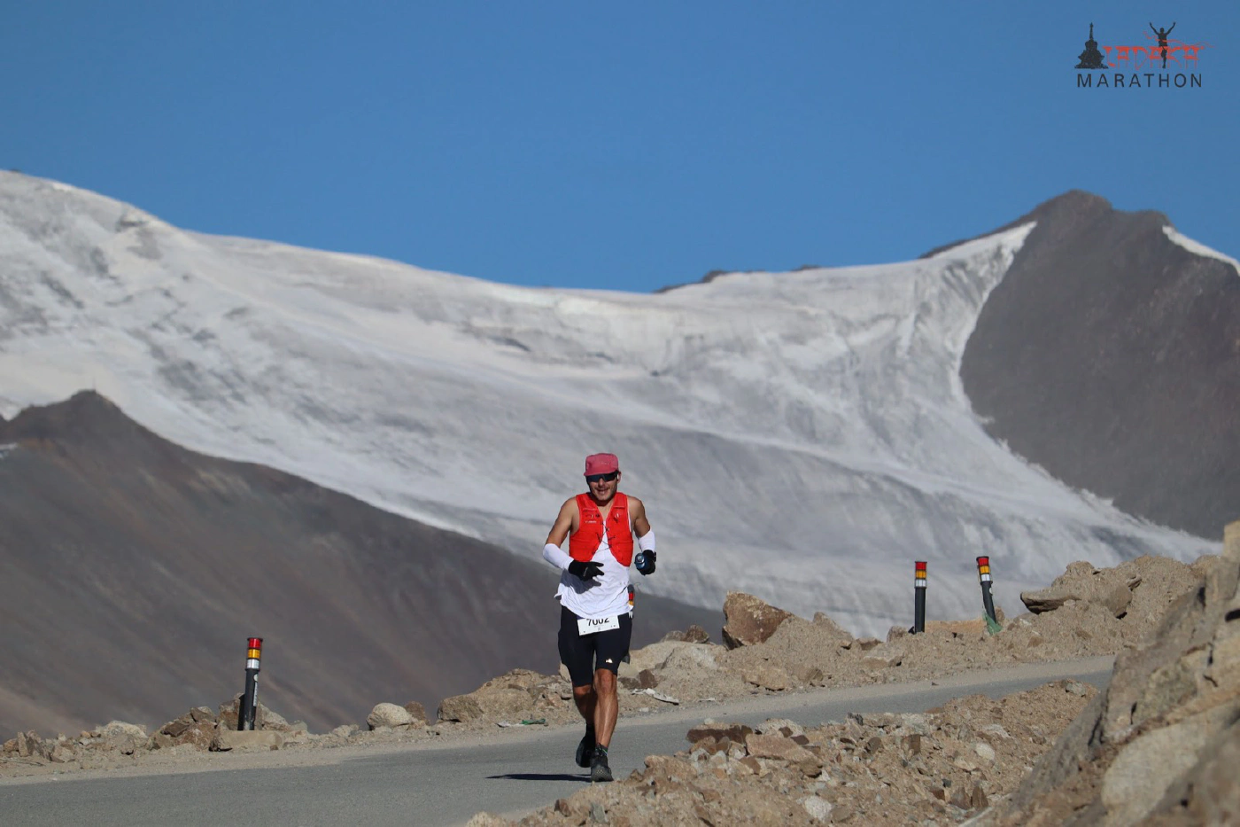 Oliver running on the mountain road in a red vest and white singlet, with a vast snow-covered peak filling the frame behind him under a deep blue sky.