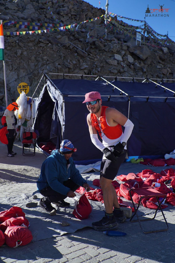 Oliver at Khardung La Top, standing beside a tent and rows of red drop bags under prayer flags, wearing a running vest over a singlet.