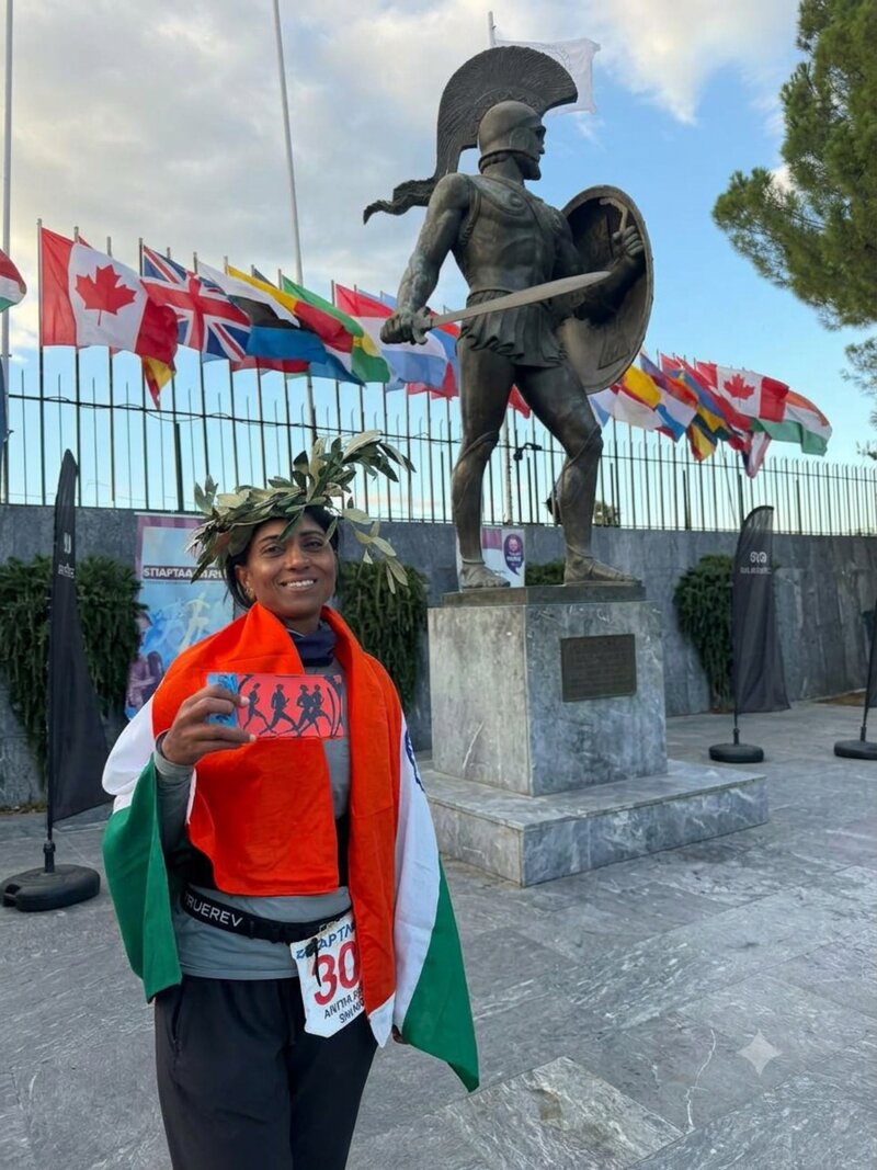 Anitha stands in front of the statue of King Leonidas in Sparta, wearing a laurel wreath and wrapped in the Indian flag, holding her finisher certificate.