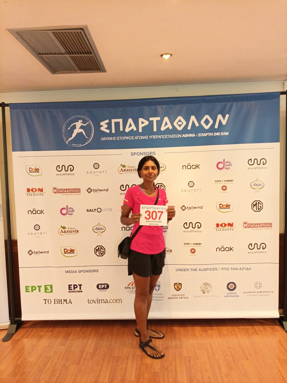 Anitha stands in front of the Spartathlon sponsor backdrop holding race number 307, wearing a pink T-shirt and black shorts.