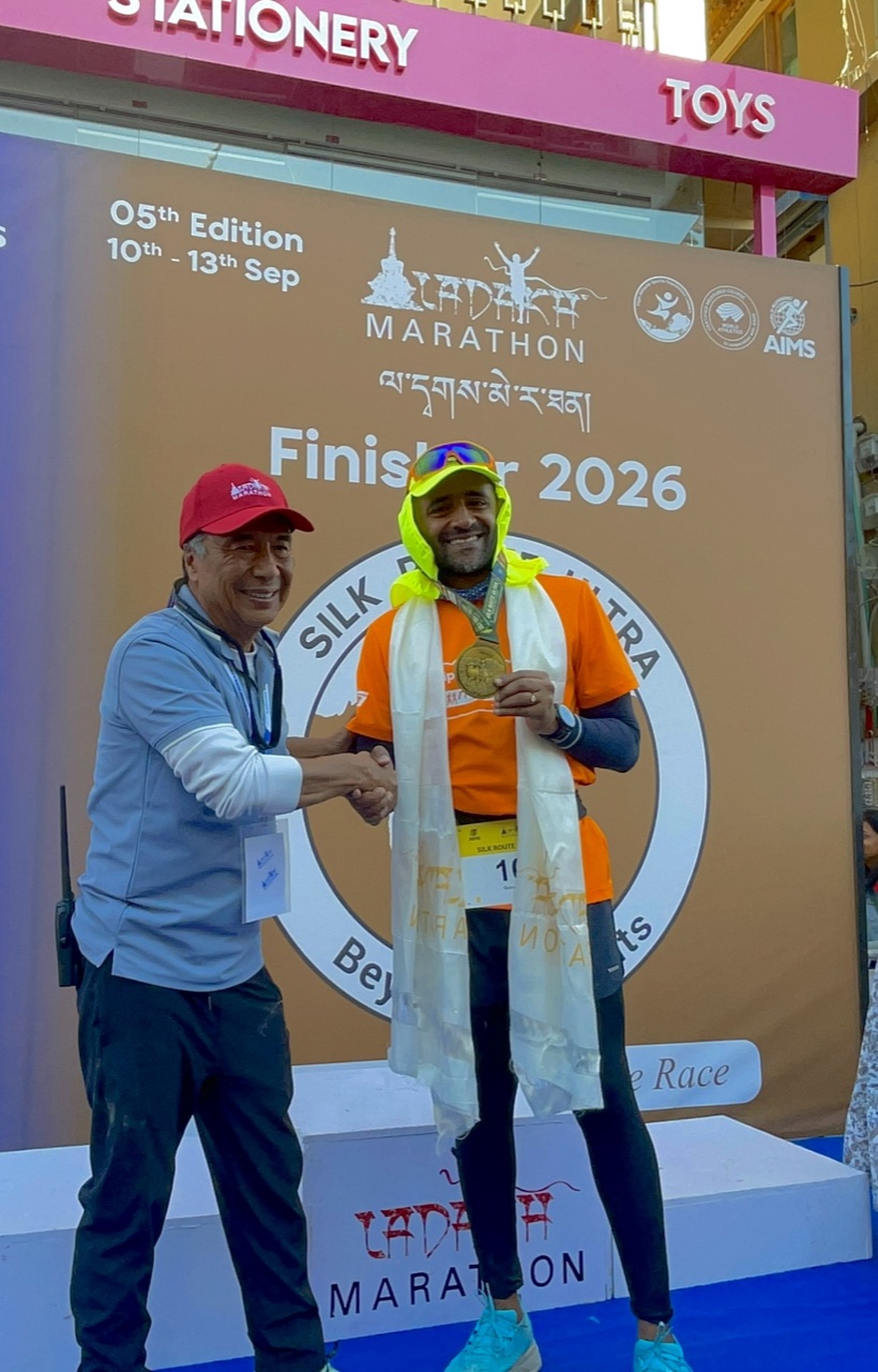 Guru, wearing a white khata scarf and his Silk Route Ultra finisher medal, shakes hands with an official in a red Ladakh Marathon cap in front of the Finisher 2026 backdrop.