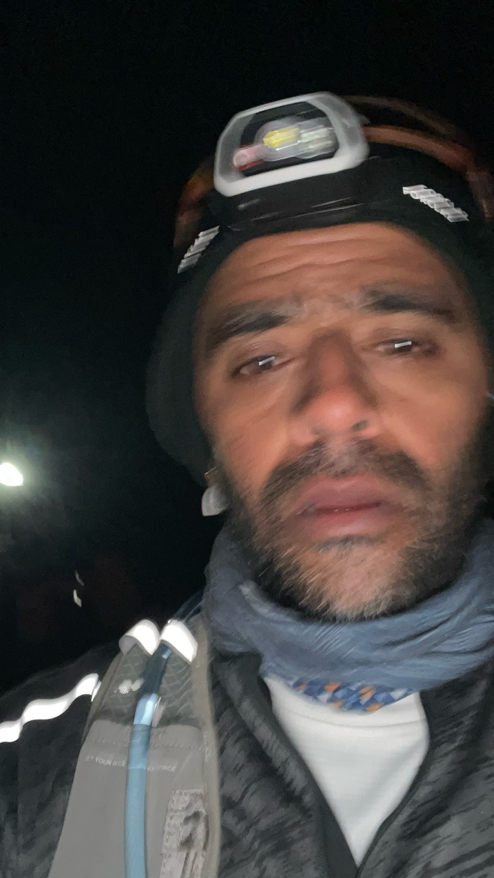 A dark, close-up selfie of Guru on the night climb, wearing a beanie and head torch, with a neck gaiter and hydration vest, looking exhausted.