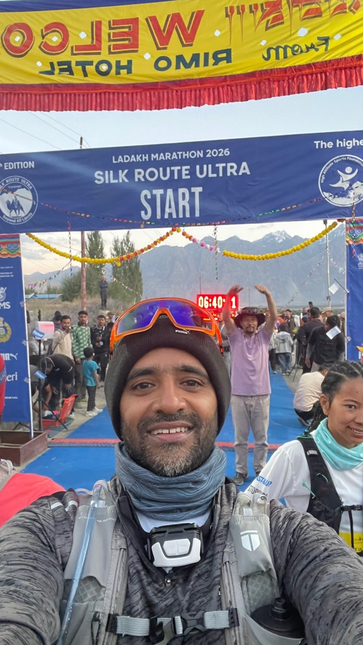 Guru takes a selfie at the Silk Route Ultra start arch in Kyagar, wearing a beanie, orange-tinted sunglasses, a neck gaiter and a head torch, with mountains and spectators behind him.