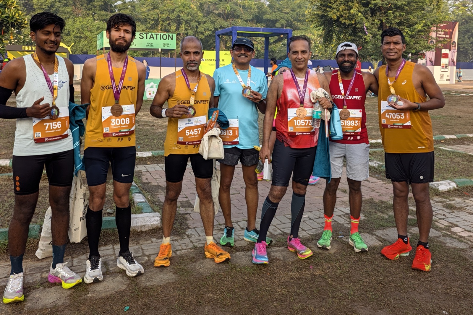 Seven runners in Geeks on Feet and other singlets stand in a row holding their finisher medals on a grass and brick path after the race.