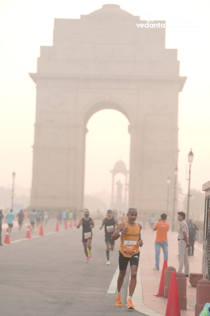 Aravind running in a yellow singlet along the avenue towards a hazy India Gate, with other runners and police behind him.
