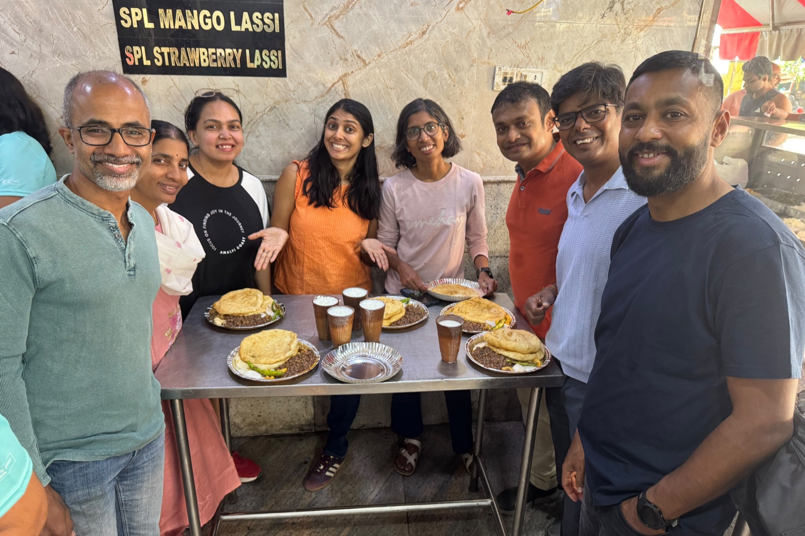 A group of friends smiling around a steel table at a lassi stall, with plates of chole bhature and glasses of lassi.