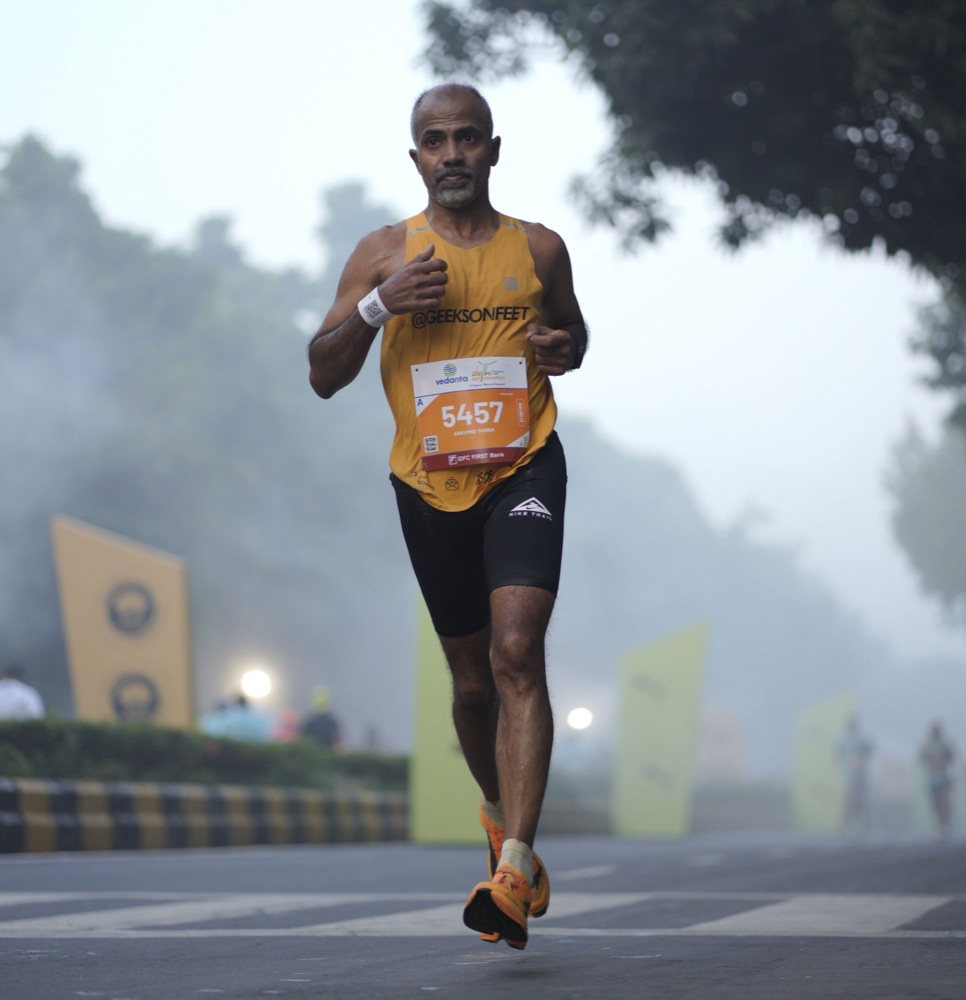 Aravind in a yellow Geeks on Feet singlet, bib 5457, running steadily through morning mist with yellow banners behind him.