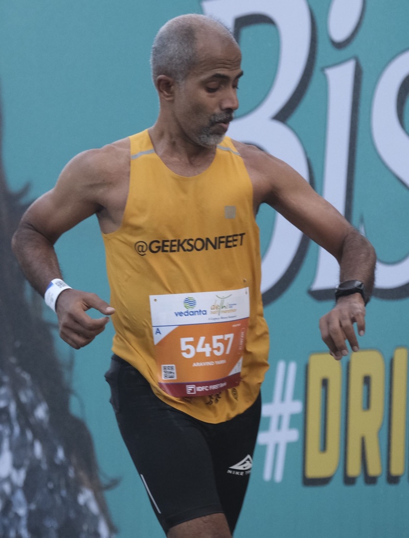 Aravind in a yellow Geeks on Feet singlet, bib 5457, glancing down at his watch while running past a teal billboard.